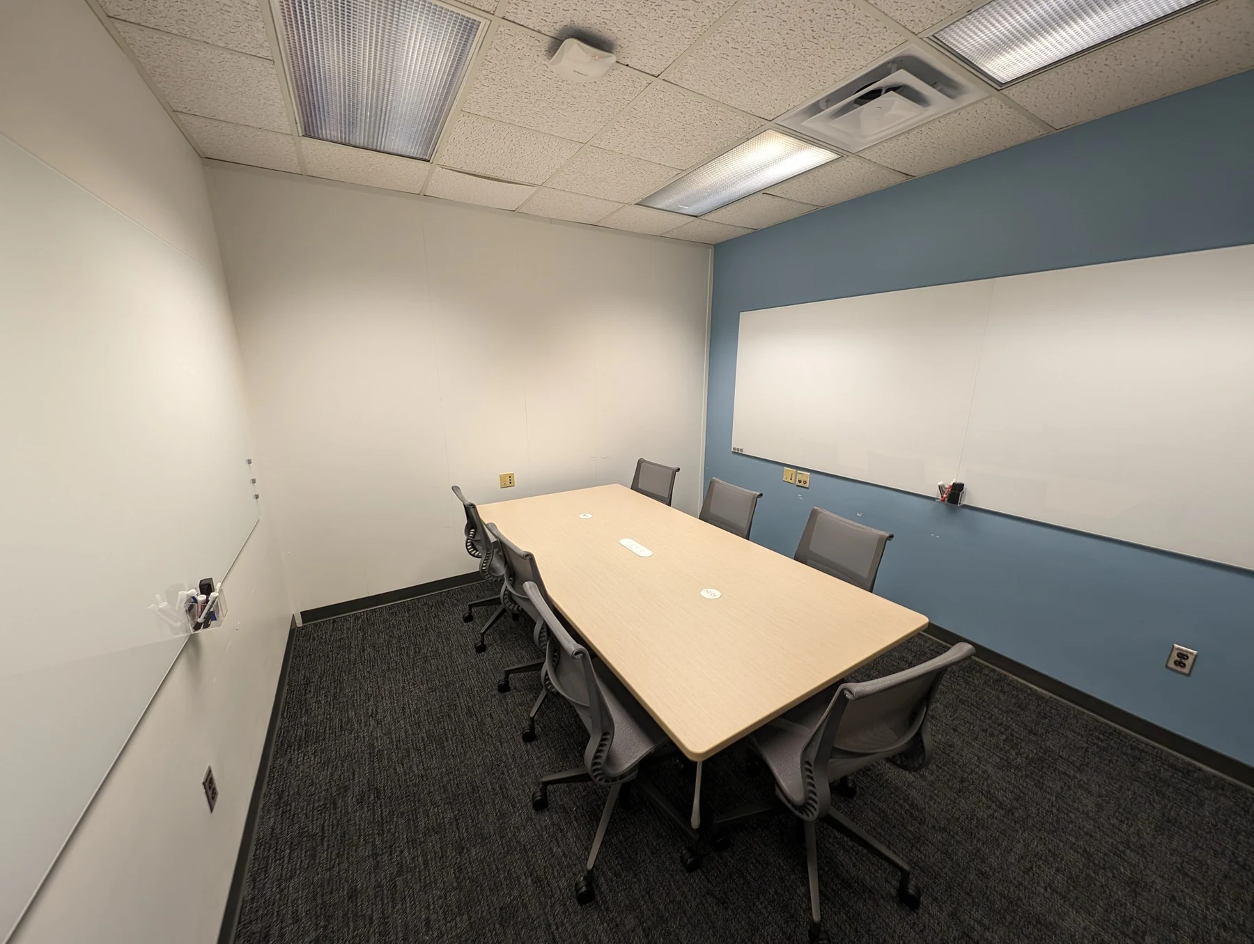 Meeting room with dark patterned carpet, a light table, gray chairs, and whiteboards on a blue accent wall