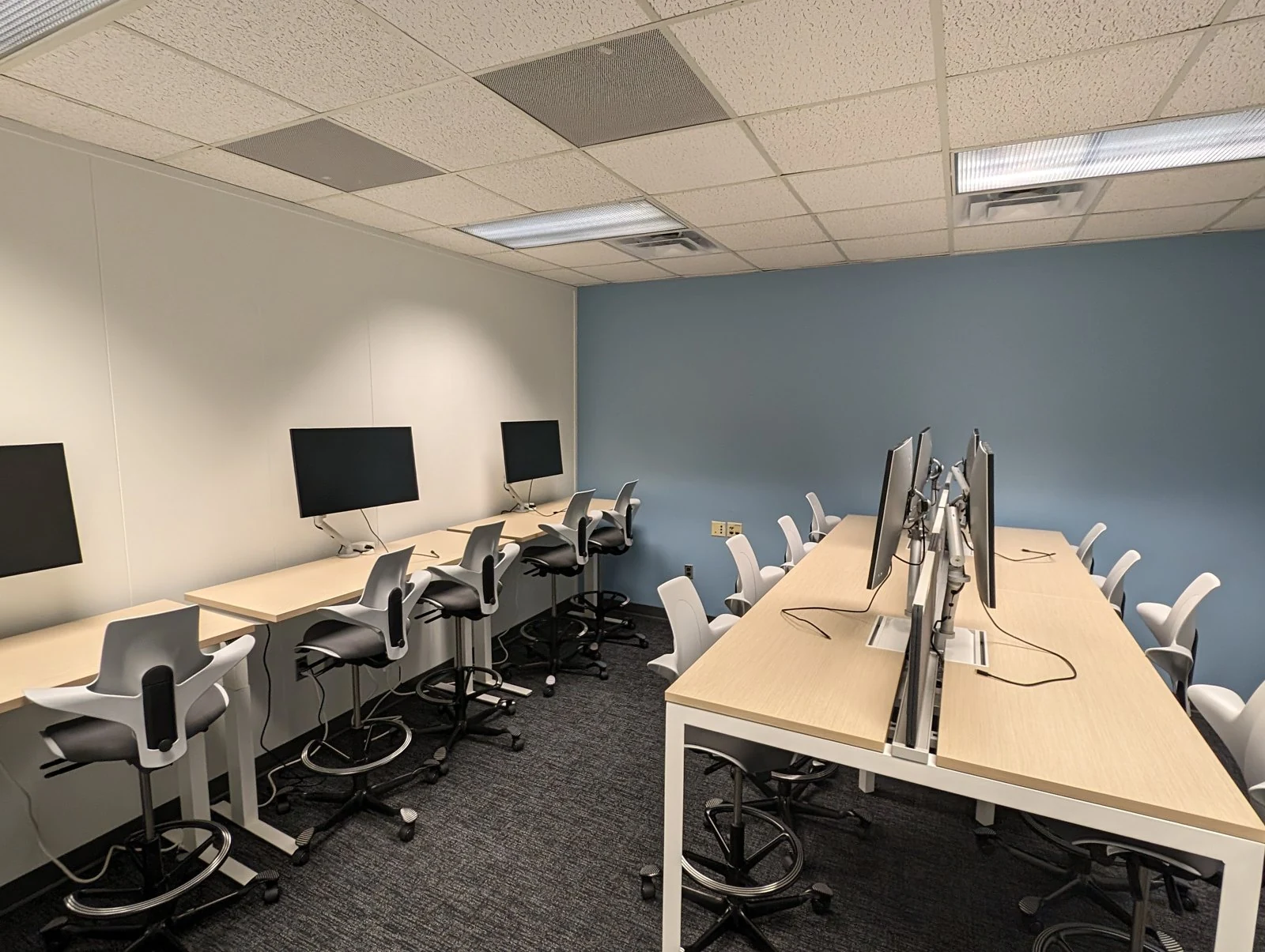 A second-phase room with two rows of white desks, monitors, monitor arms and ergonomic chairs
